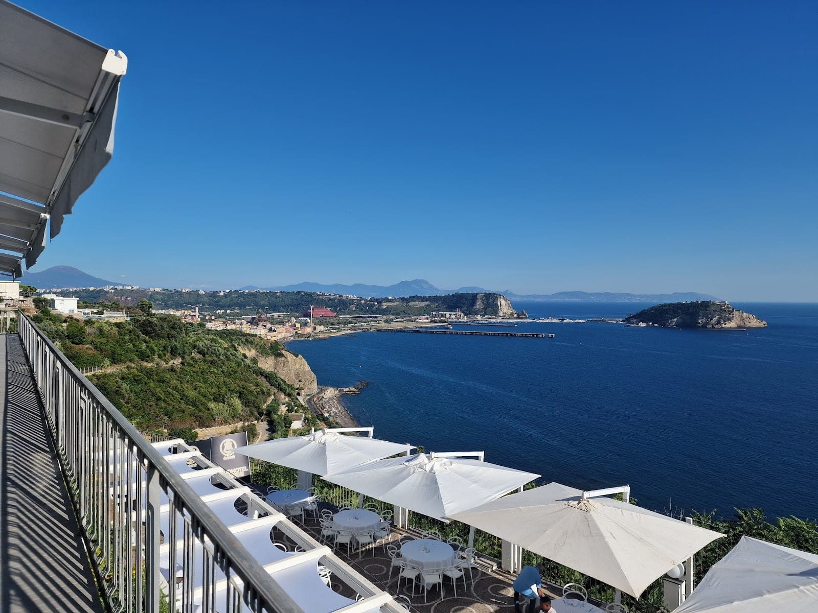 Terrace with tables and umbrellas overlooking the sea and a distant town.