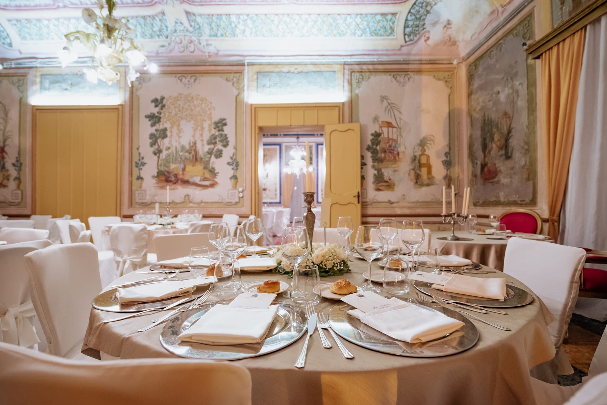 A formal dining table set with white linens and silverware in a room with painted murals.