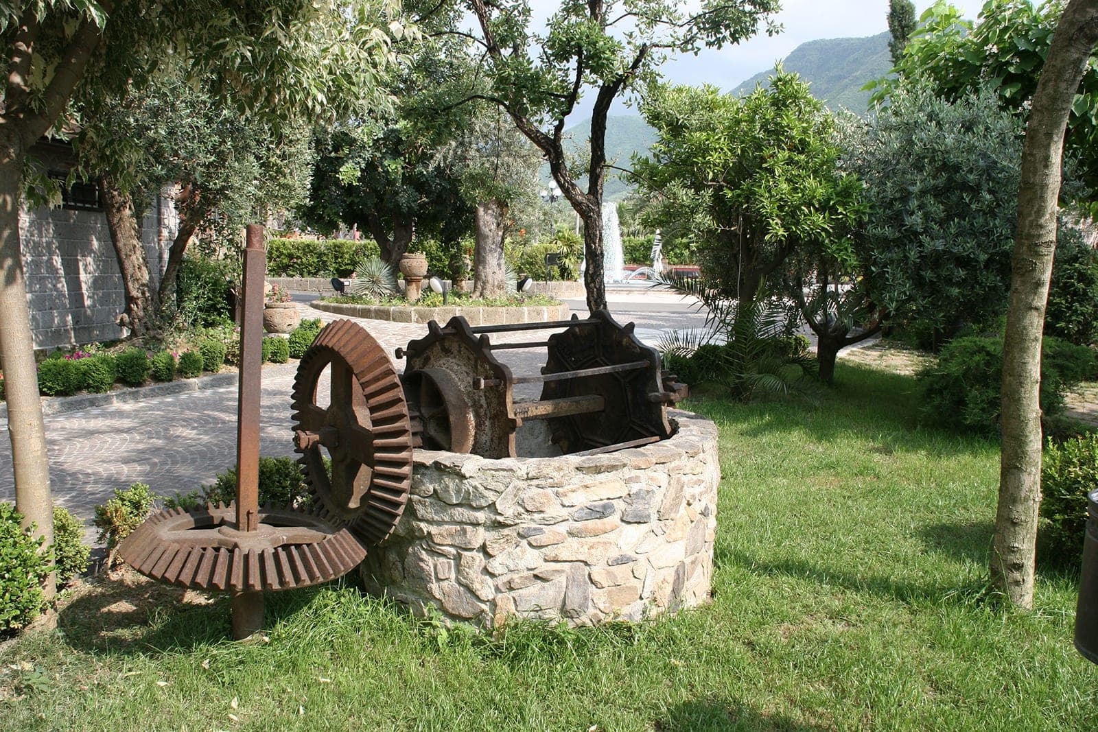 A stone well with large rusty gears in a garden with trees and a fountain in the background.