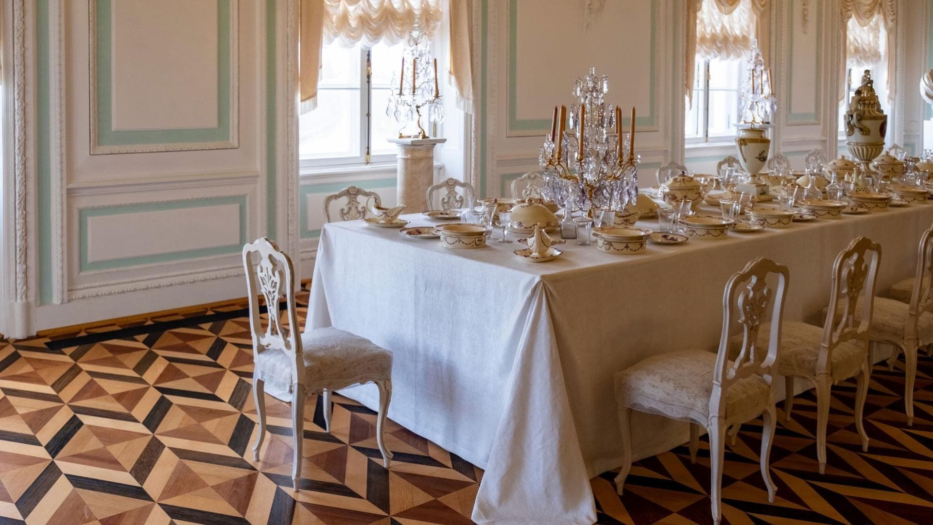 A long dining table set with fine china and crystal in a room with ornate walls and a patterned floor.
