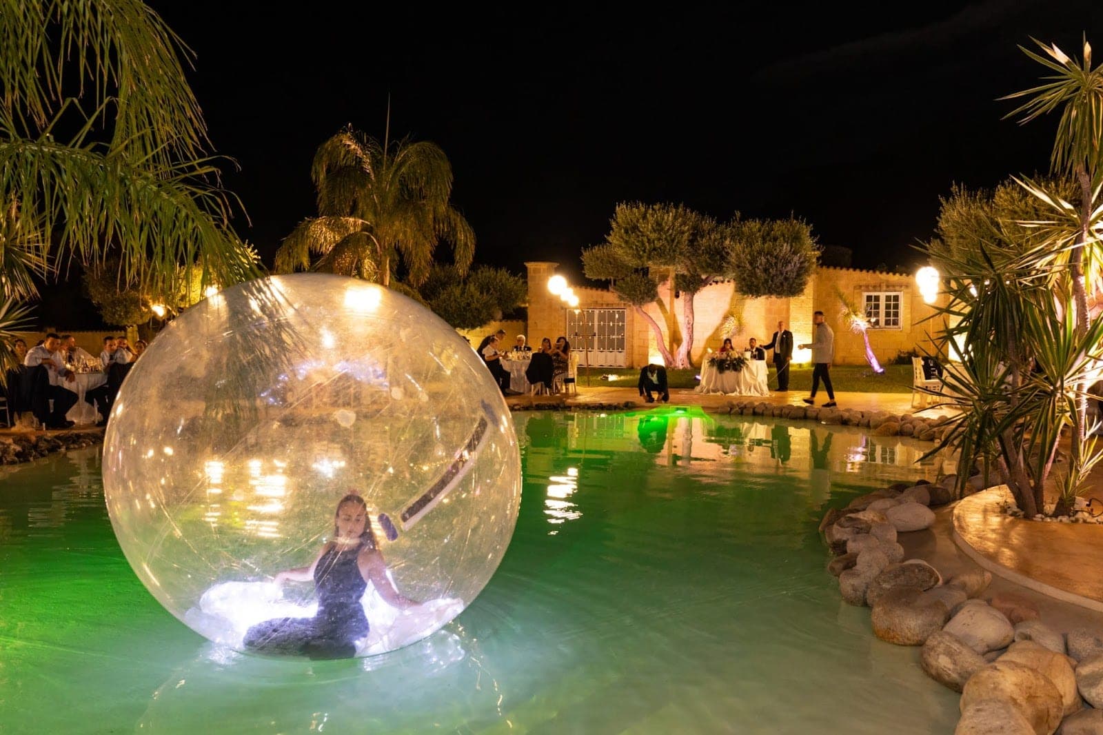 A wedding reception at night with tables around a pool and a person in a large ball.