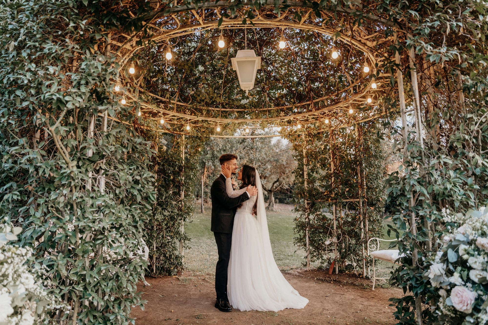 A couple embraces under a floral archway decorated with string lights.