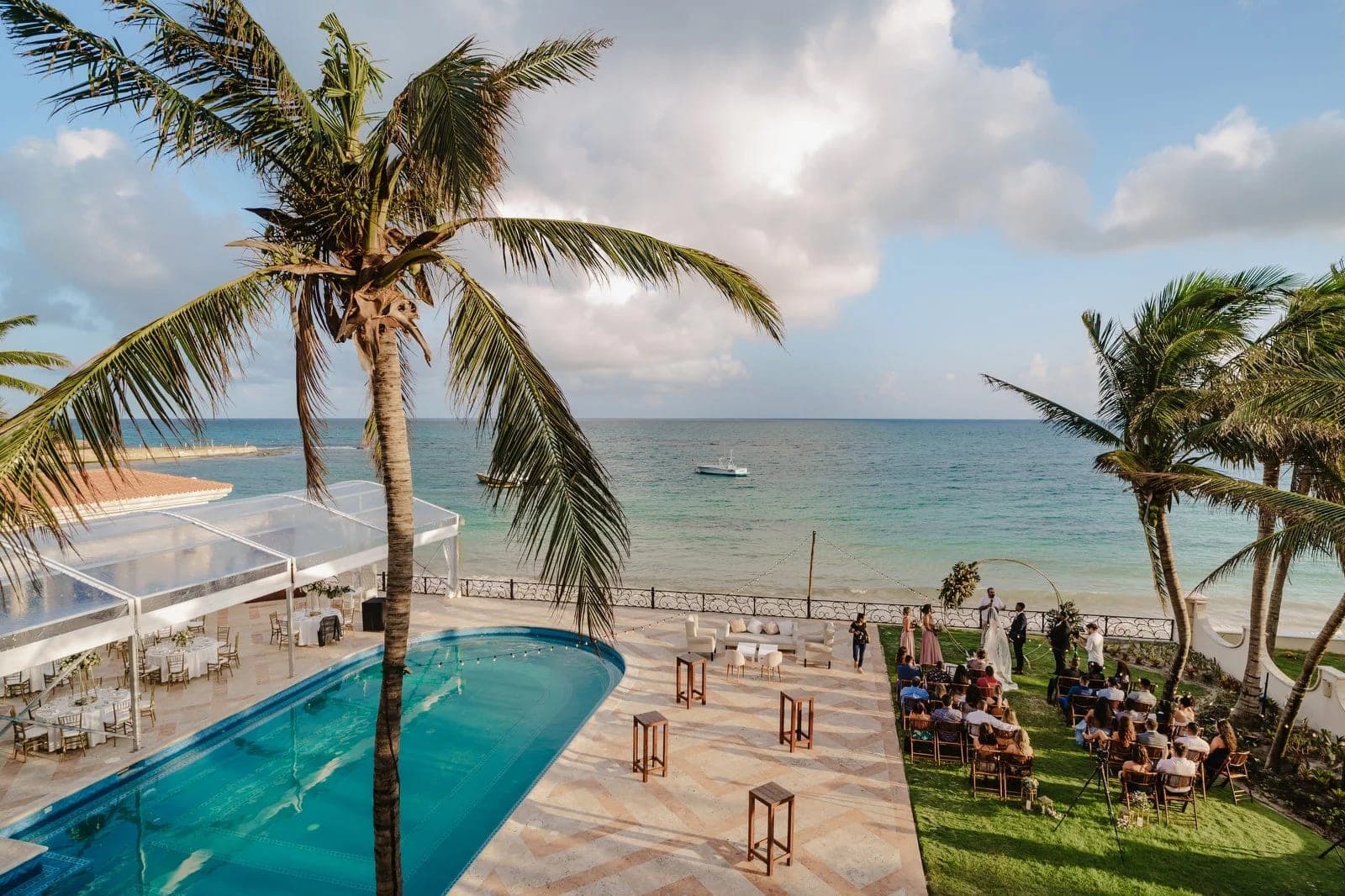 Wedding ceremony setup on a lawn overlooking the ocean with palm trees and a pool.