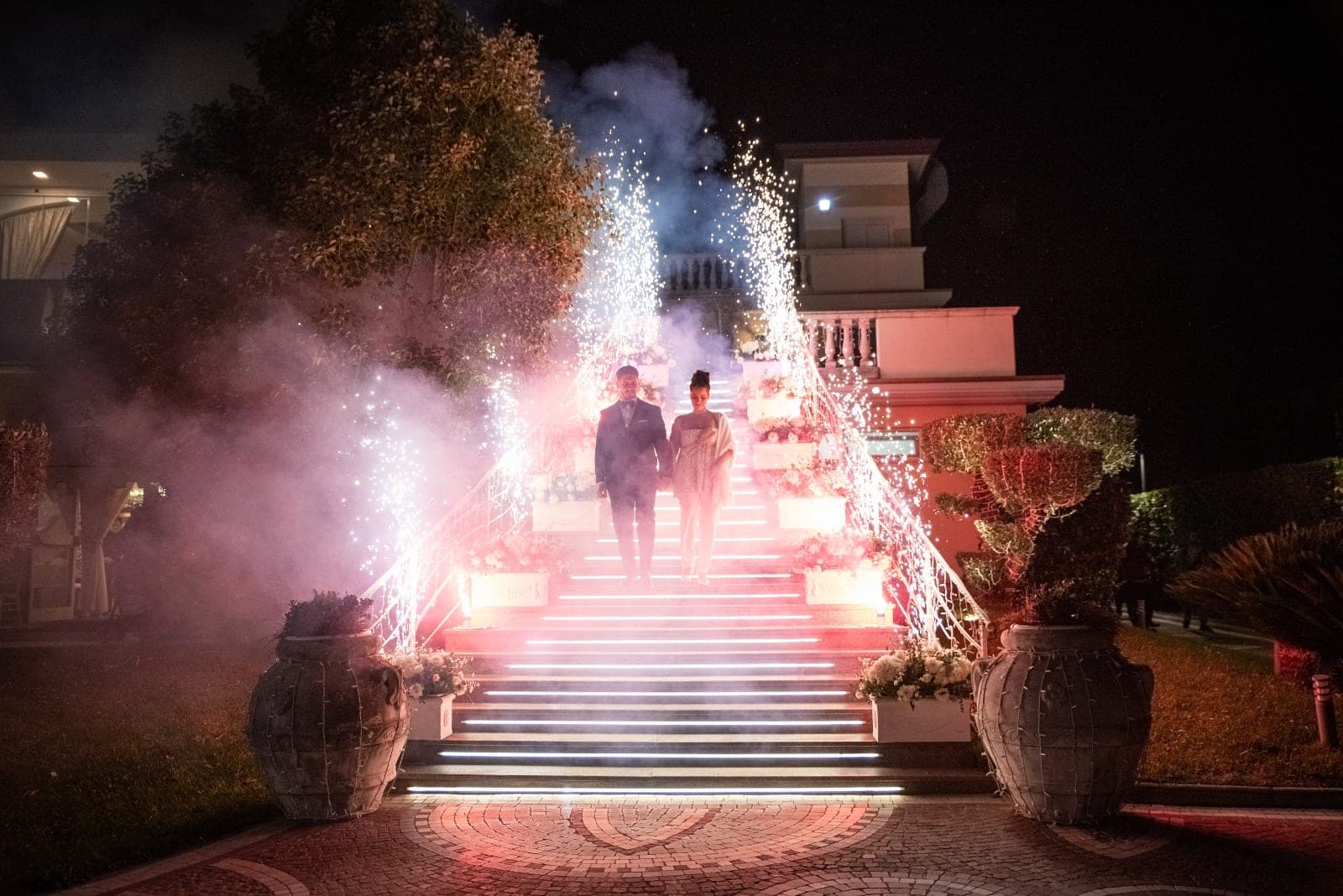 A couple walks down a staircase illuminated by fireworks and lights.