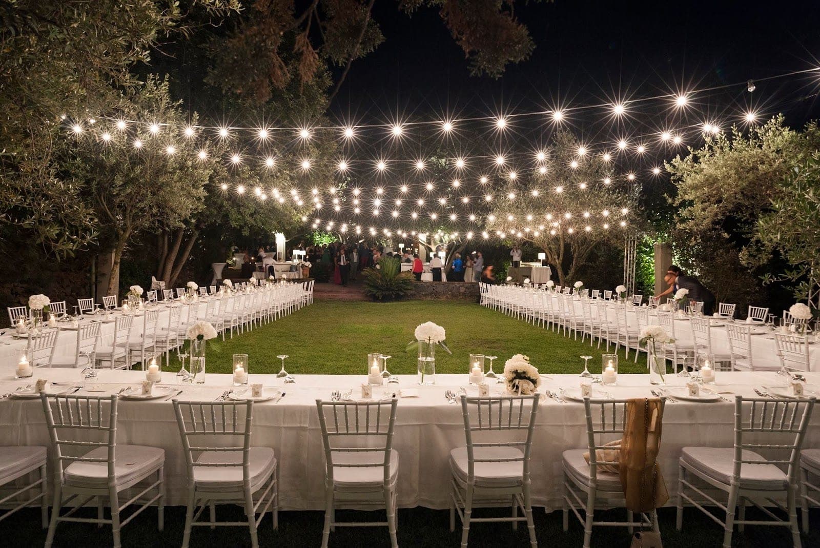 Outdoor reception tables set for a wedding dinner under string lights at night.