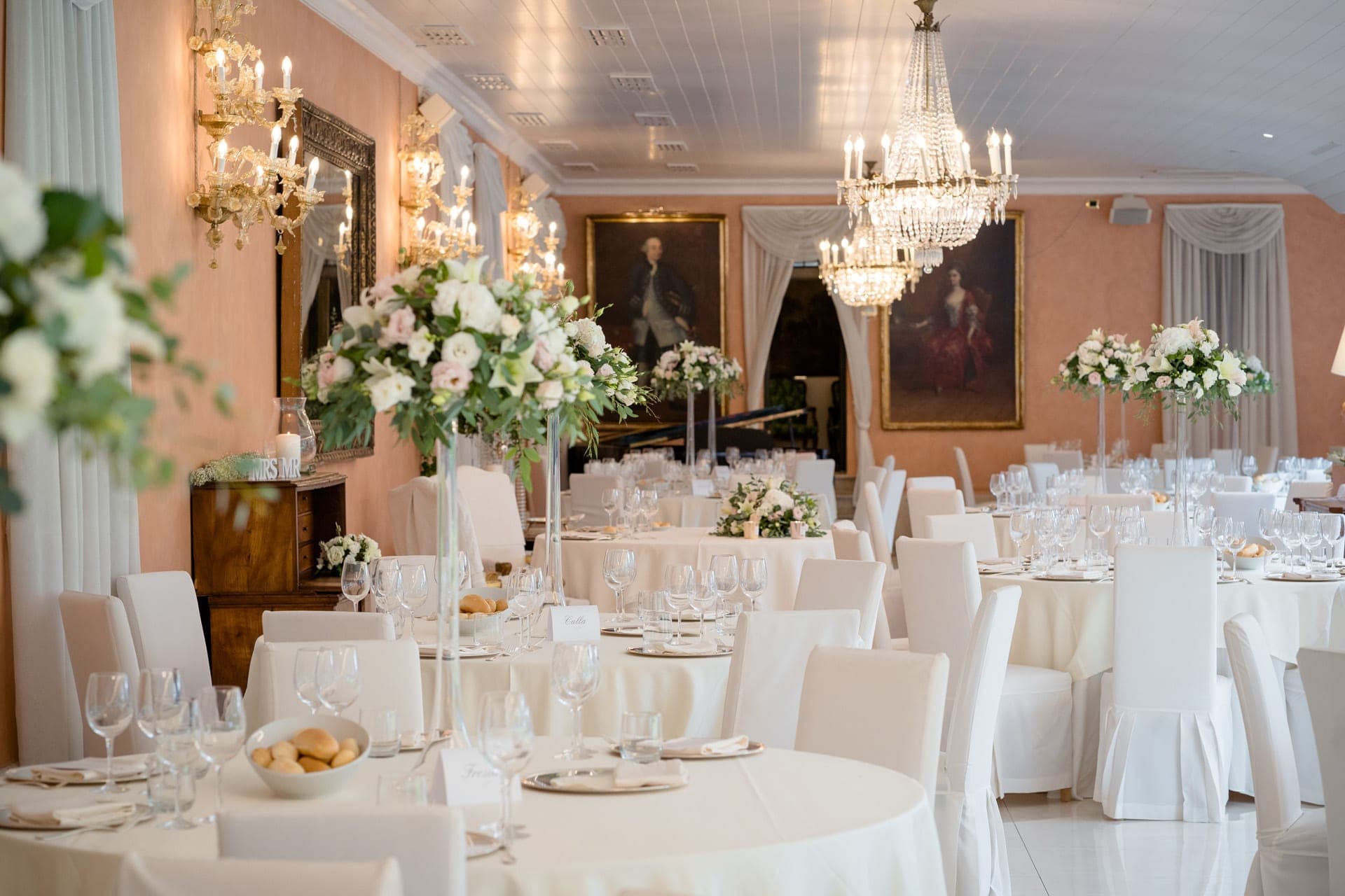 A wedding reception room with round tables set for dinner, chandeliers, and floral centerpieces.