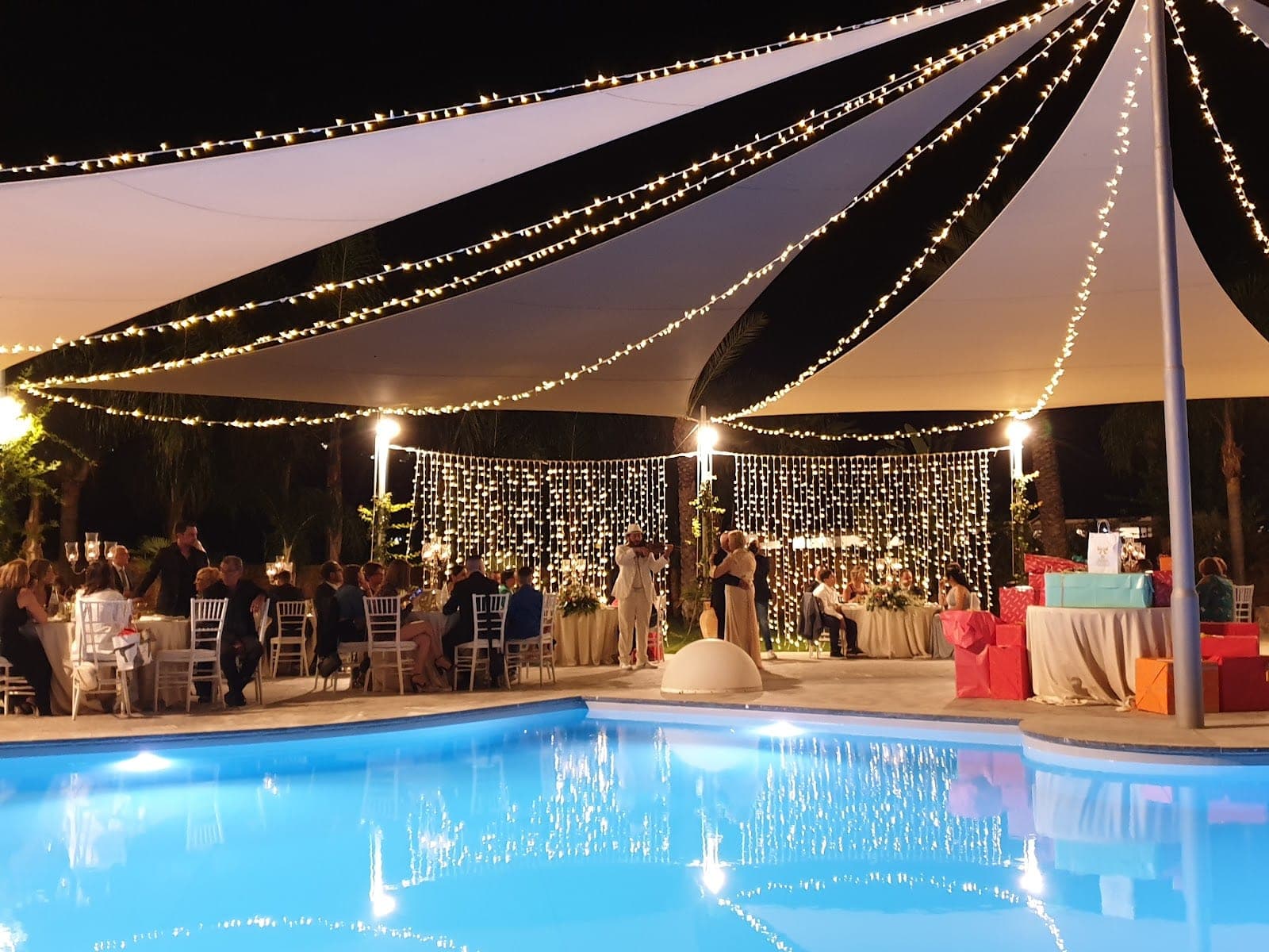 A wedding reception is set up outdoors at night with tables, chairs, a pool, and string lights.