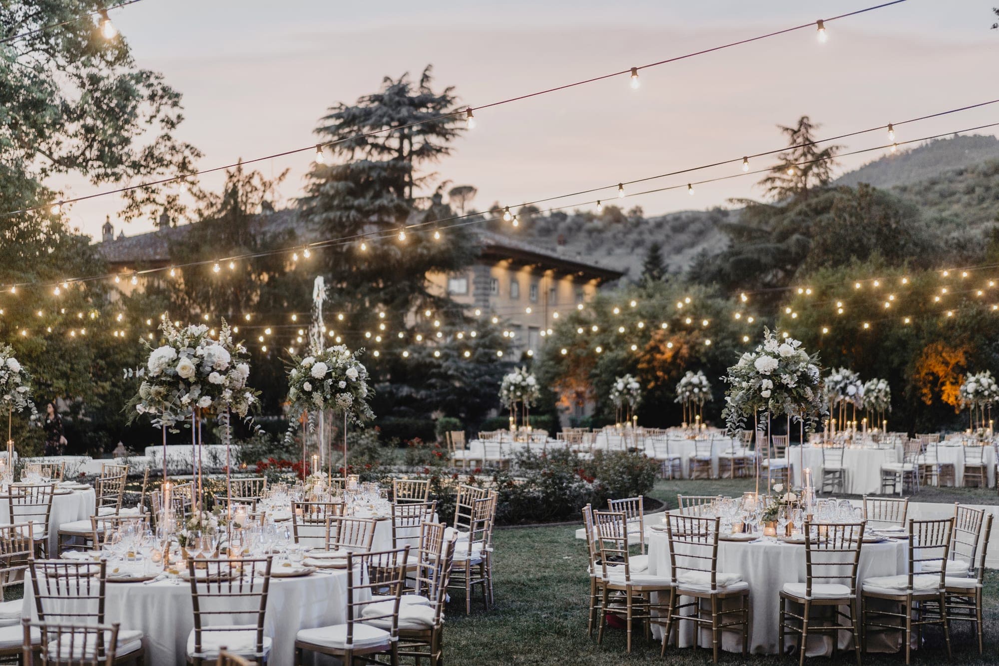 Outdoor reception tables set with white linens and gold chairs under string lights at dusk.