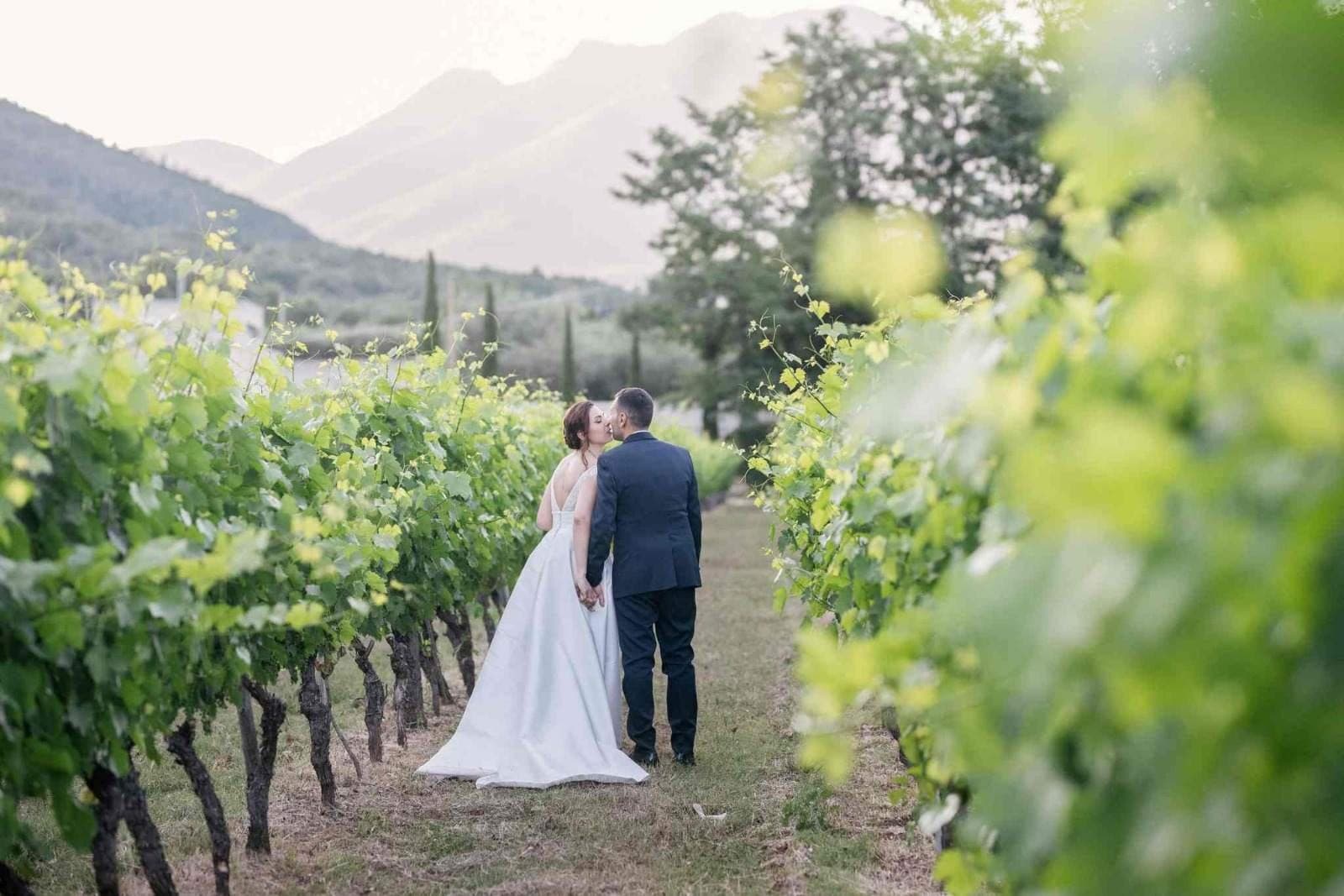 A couple kisses in a vineyard with mountains in the background.