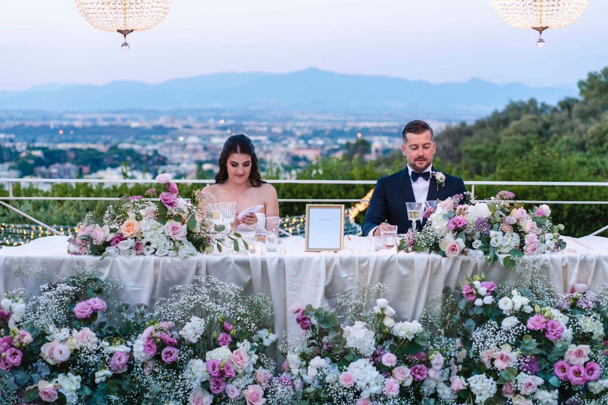 A bride and groom sit at a flower-adorned table overlooking a city at dusk.