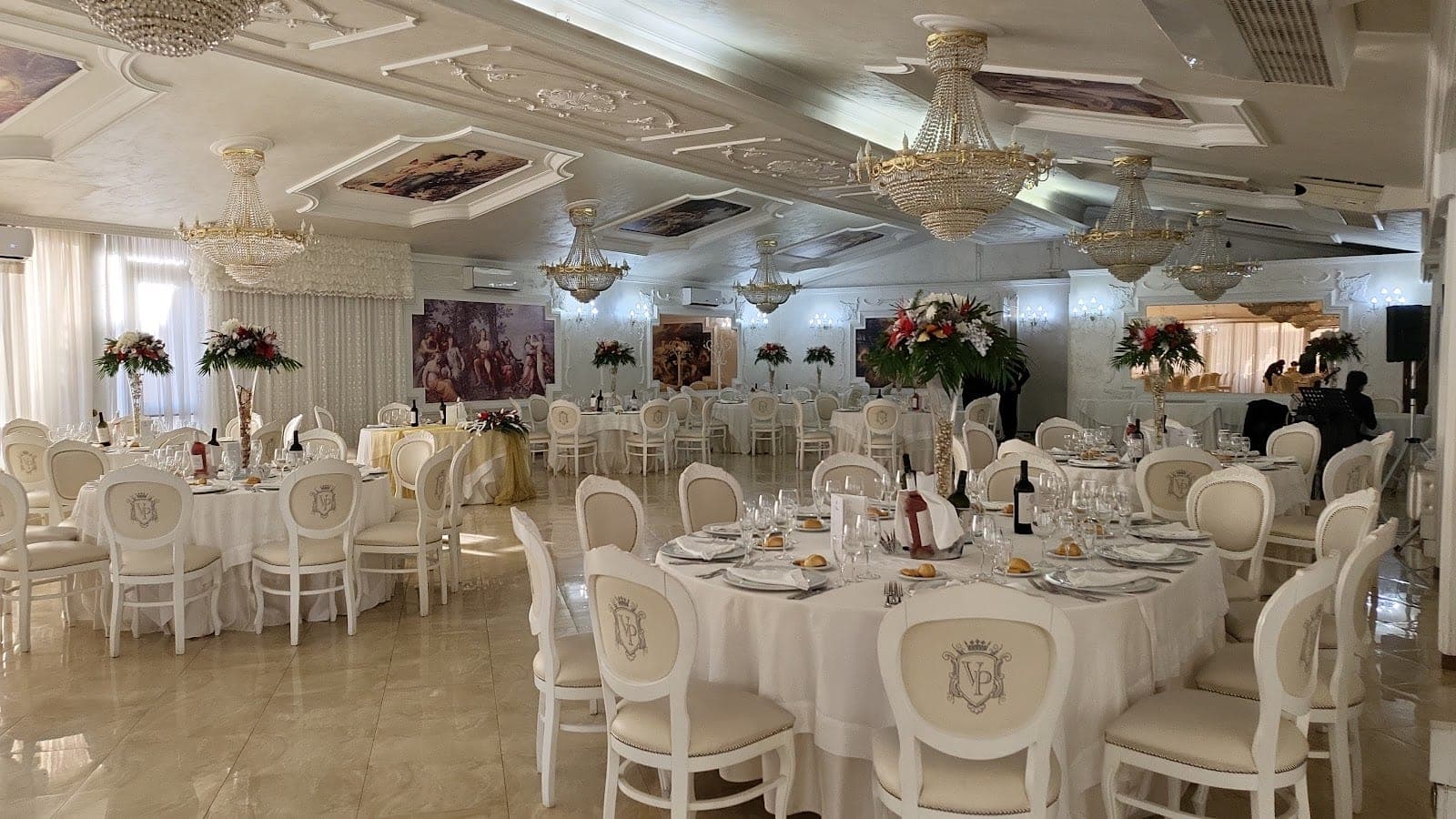 A reception room with round tables set for a wedding, chandeliers, and floral centerpieces.