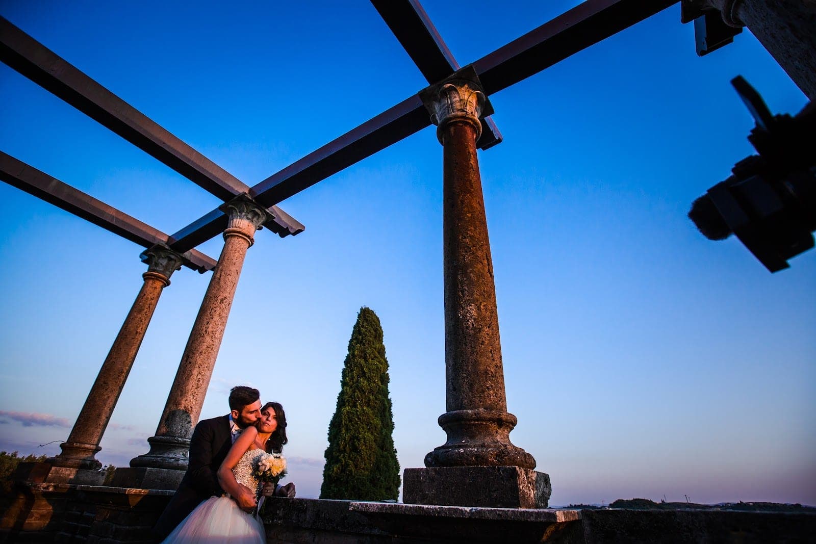 A couple embraces on a terrace with ancient columns and a pergola.