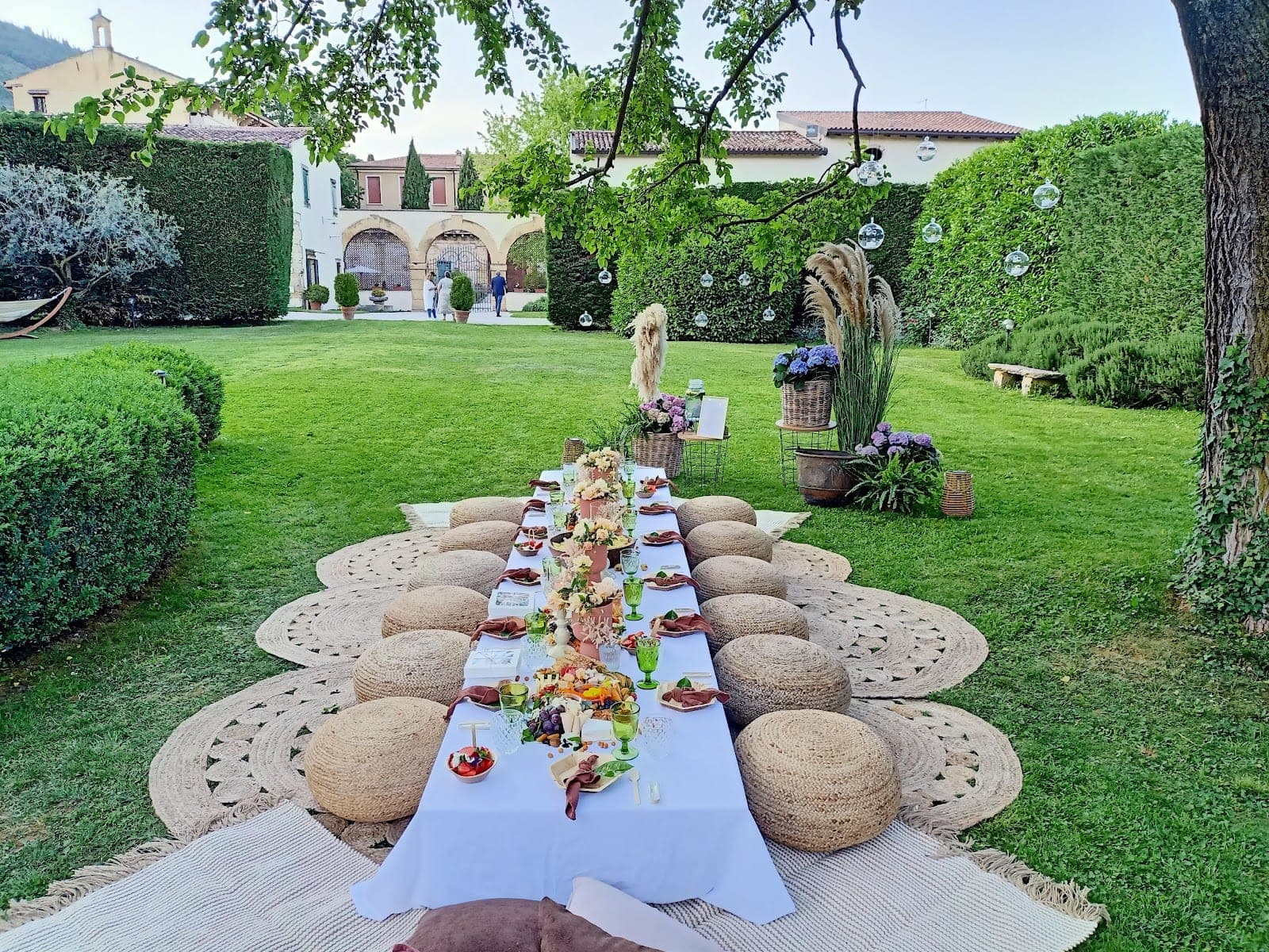 Outdoor reception dining table set on a lawn with cushions and a villa in the background.