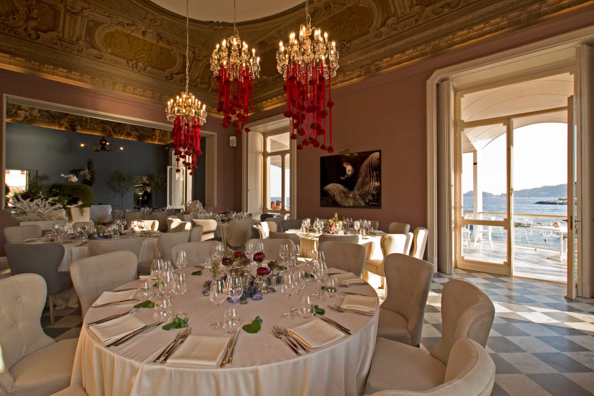 A formal dining room with round tables set for a wedding reception, overlooking the sea.