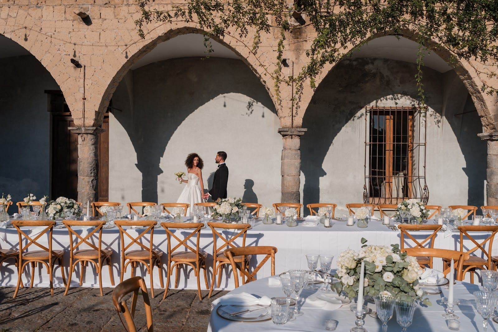 Wedding reception tables set outdoors under stone arches with a bride and groom.