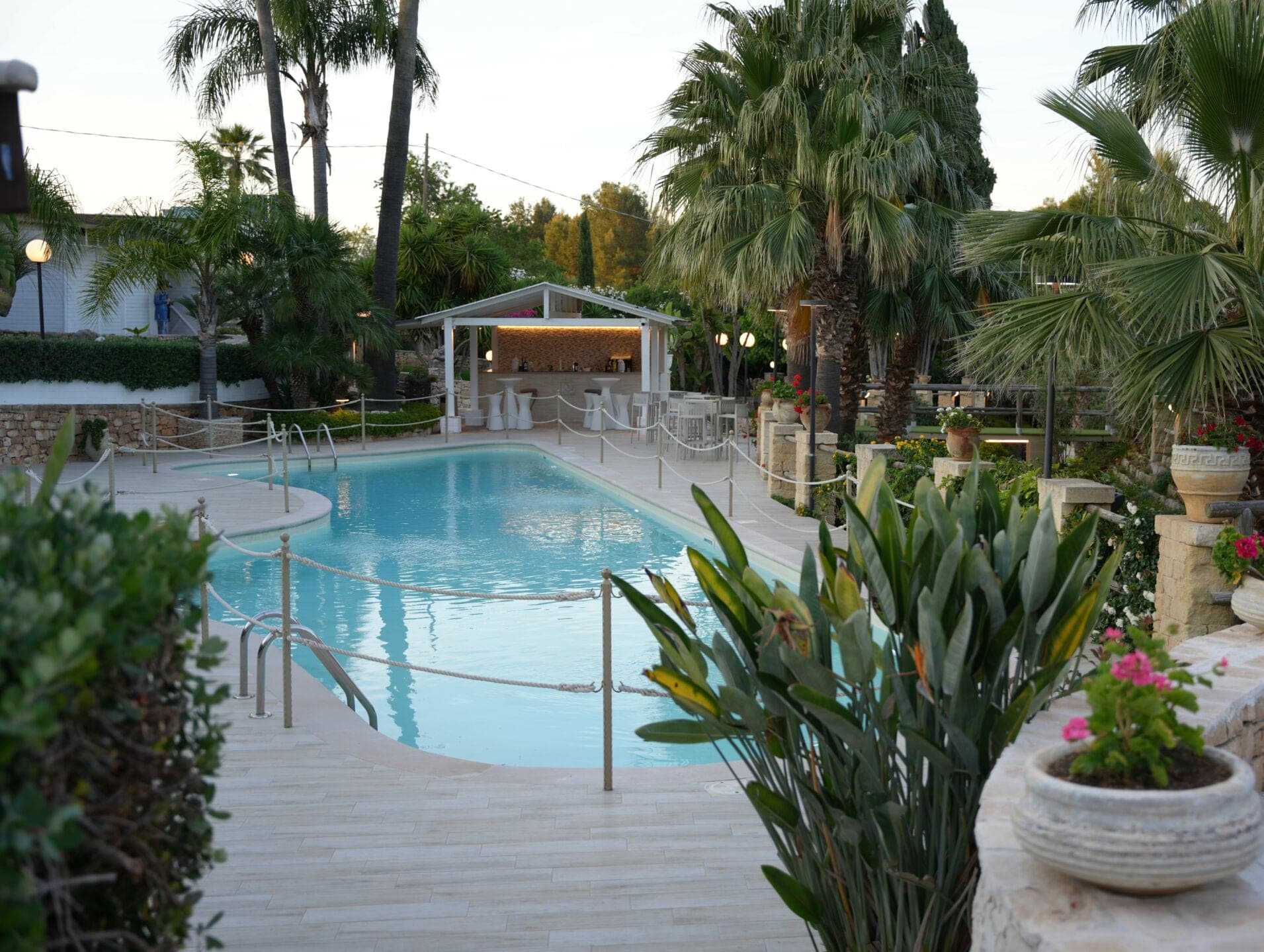 Swimming pool and bar area at dusk with palm trees and plants.