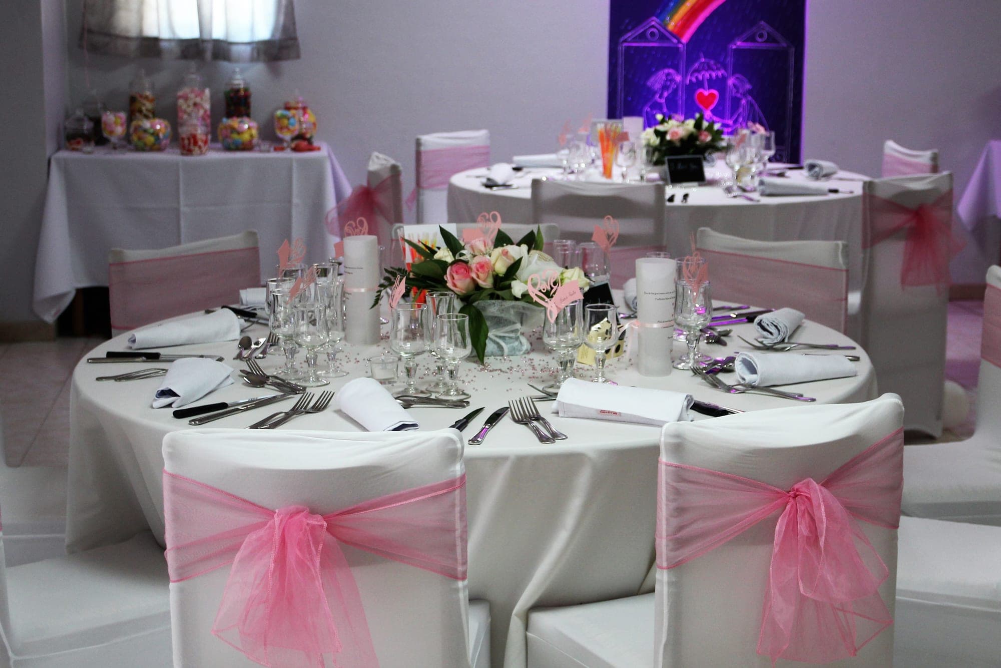 A wedding reception table is set with white linens, pink sashes on chairs, and a floral centerpiece.