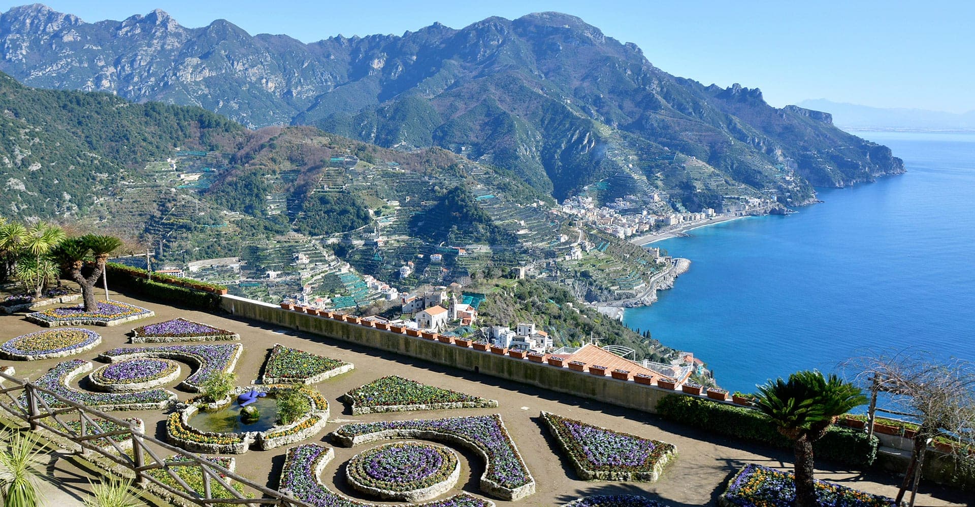 Terraced gardens with floral displays overlook a coastal town and mountains.
