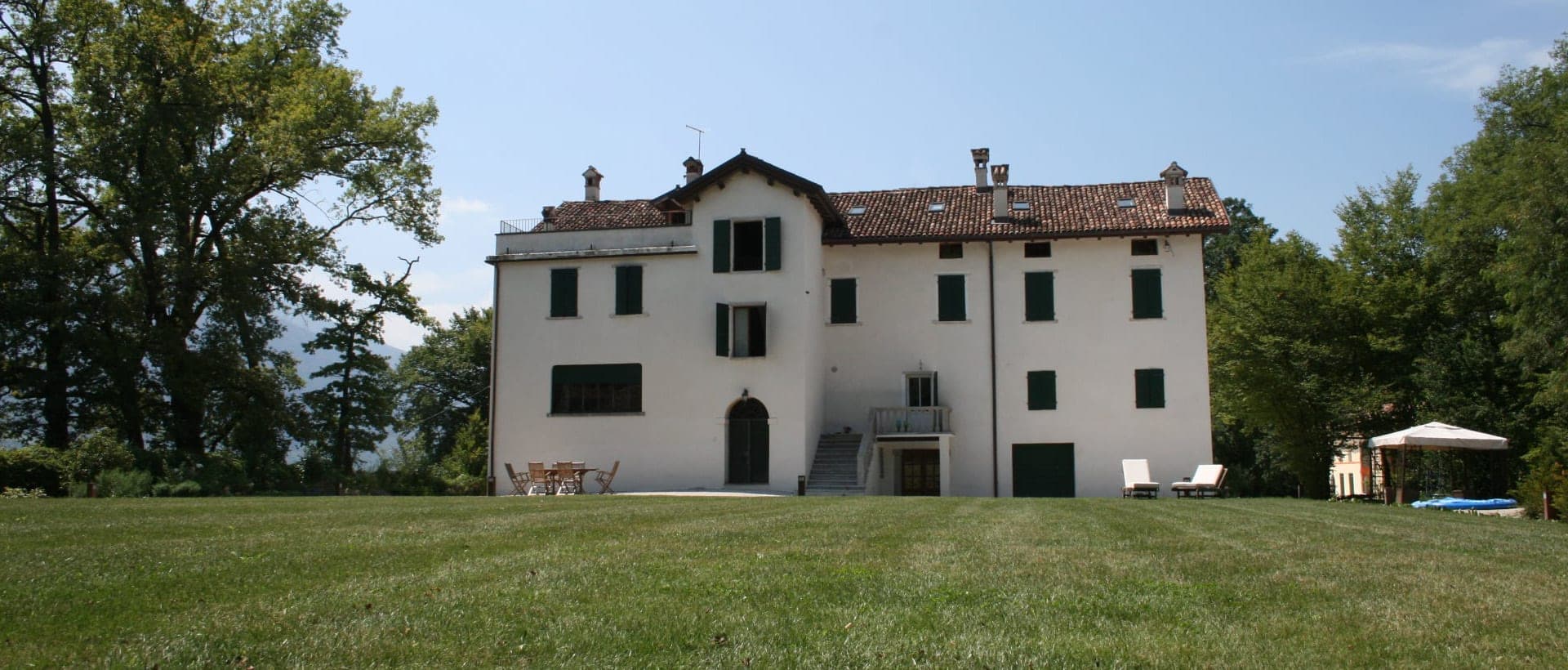 Exterior of a white villa with green shutters surrounded by trees and a lawn.
