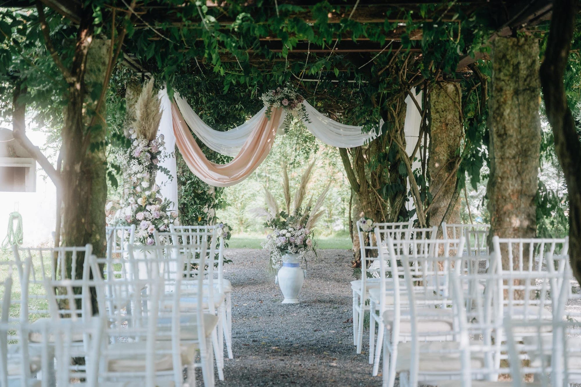 Wedding ceremony setup with white chairs and floral decorations under a leafy canopy.
