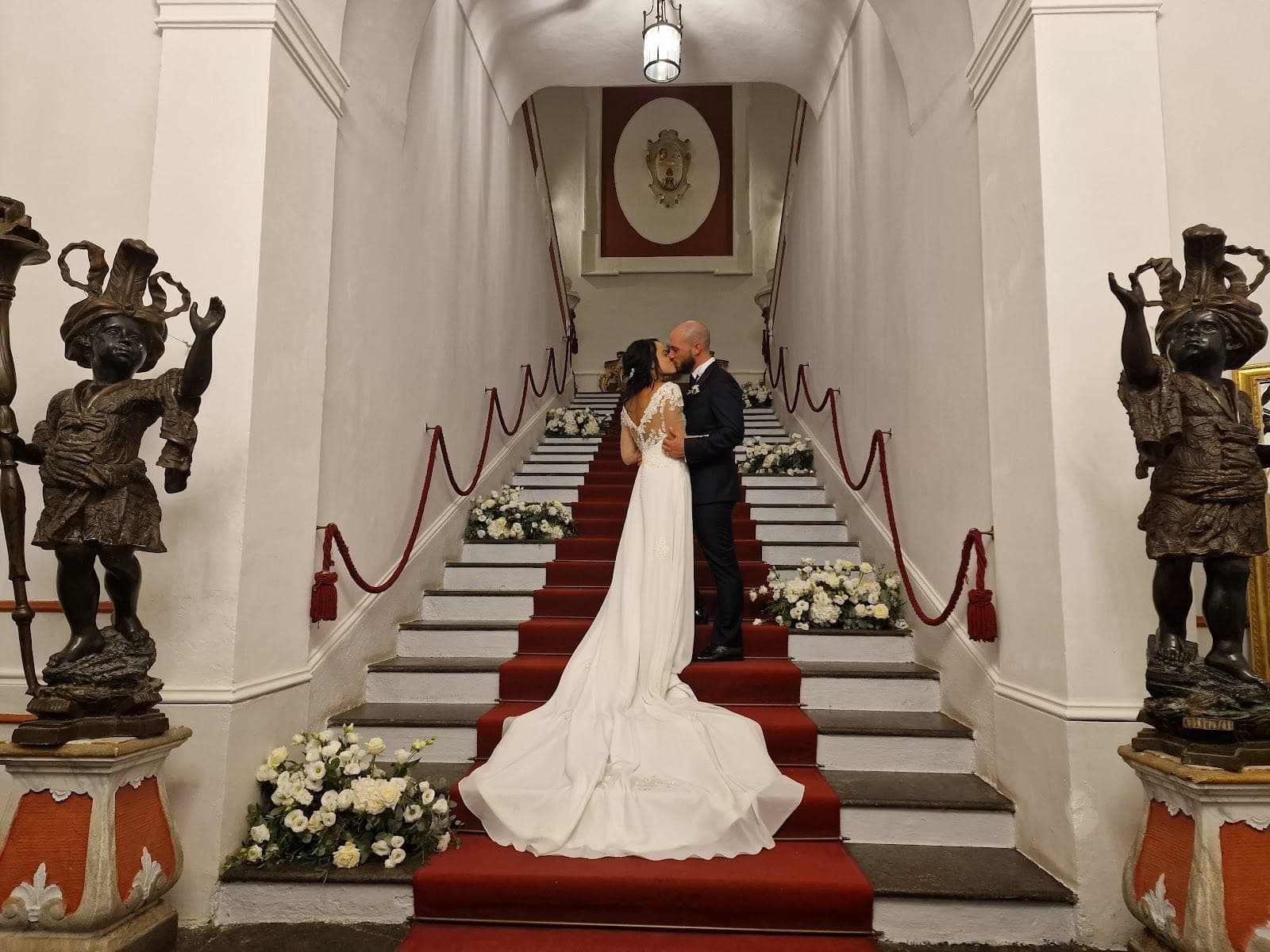 A bride and groom kiss on a red carpeted staircase with white flowers.