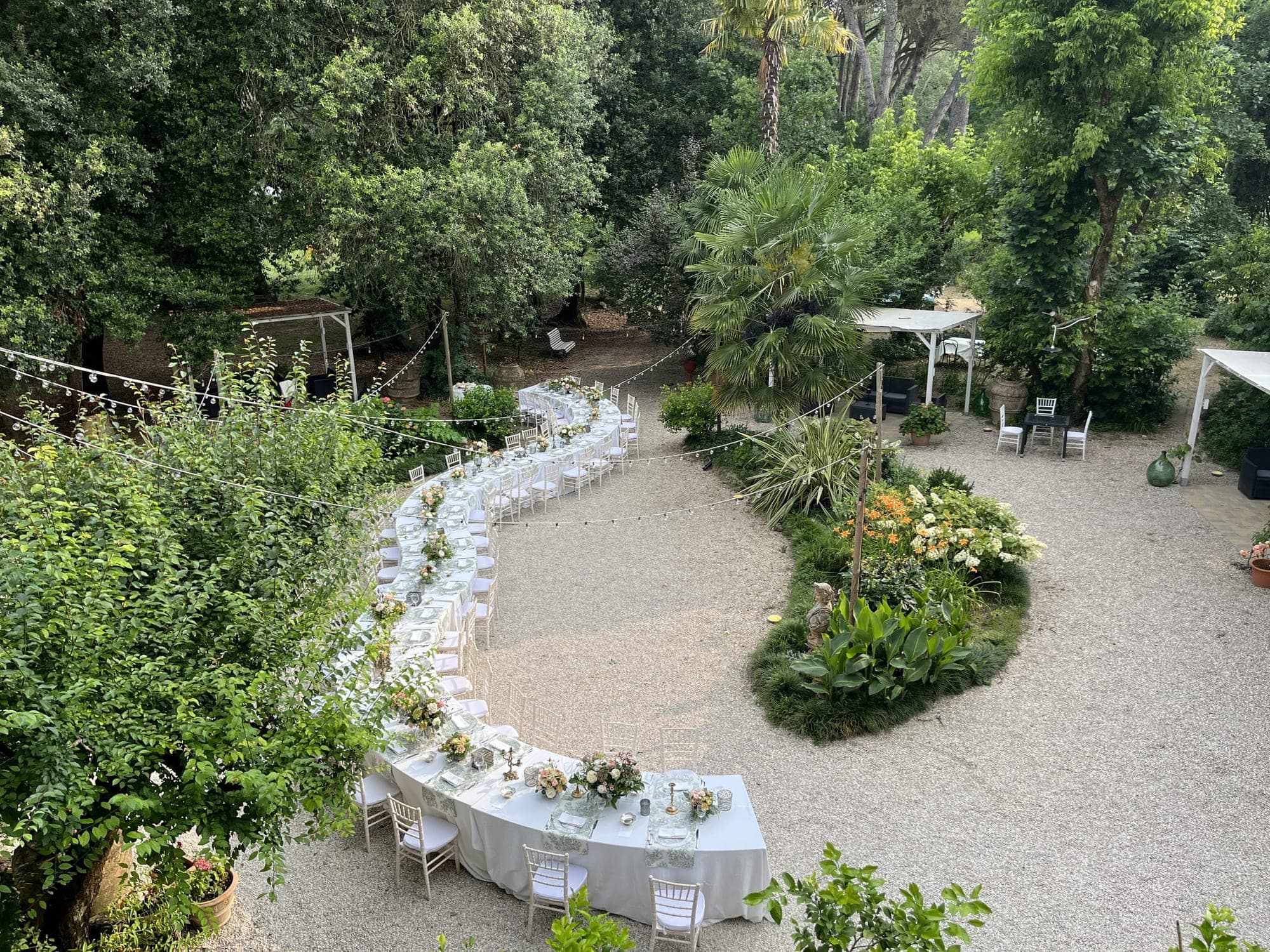 A long, curved table is set for a wedding reception in a garden with trees and plants.