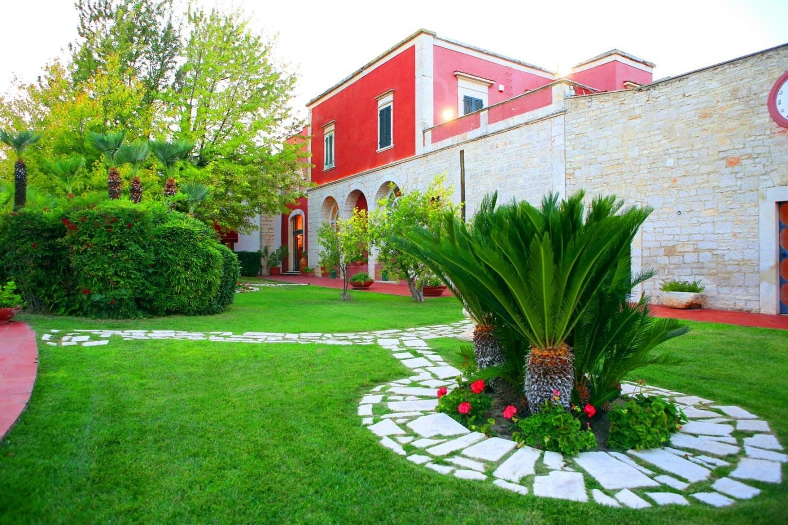 A red building with arched windows and a manicured lawn with stone pathways and palm trees.