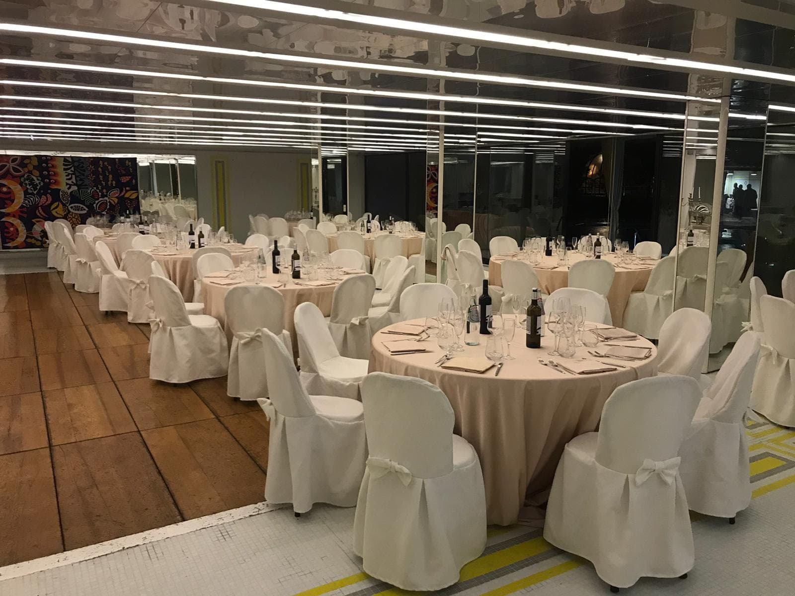 Round tables with white chair covers are set for a reception in a room with mirrored walls.