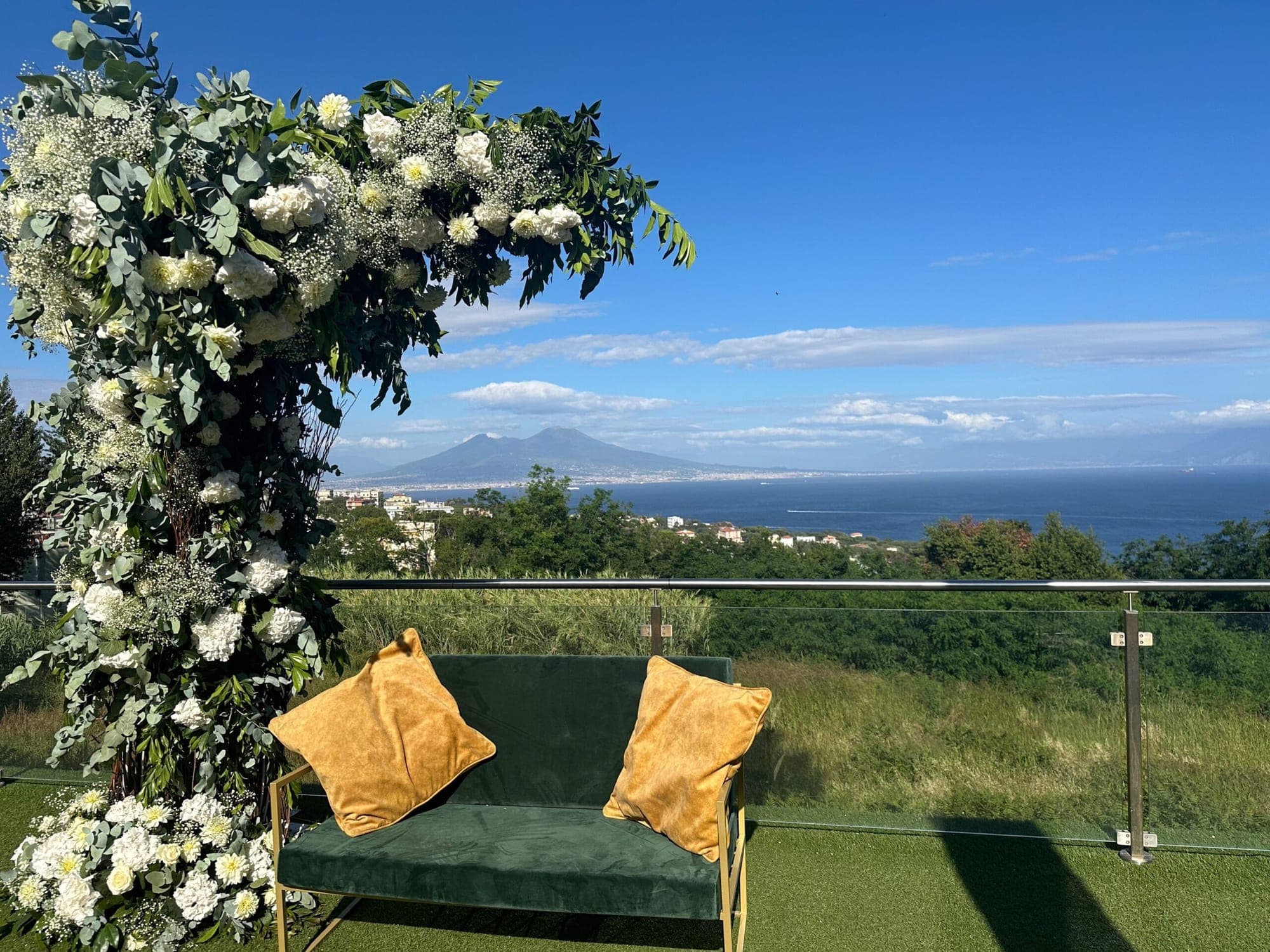 A floral arch and green bench overlook the sea and mountains under a blue sky.