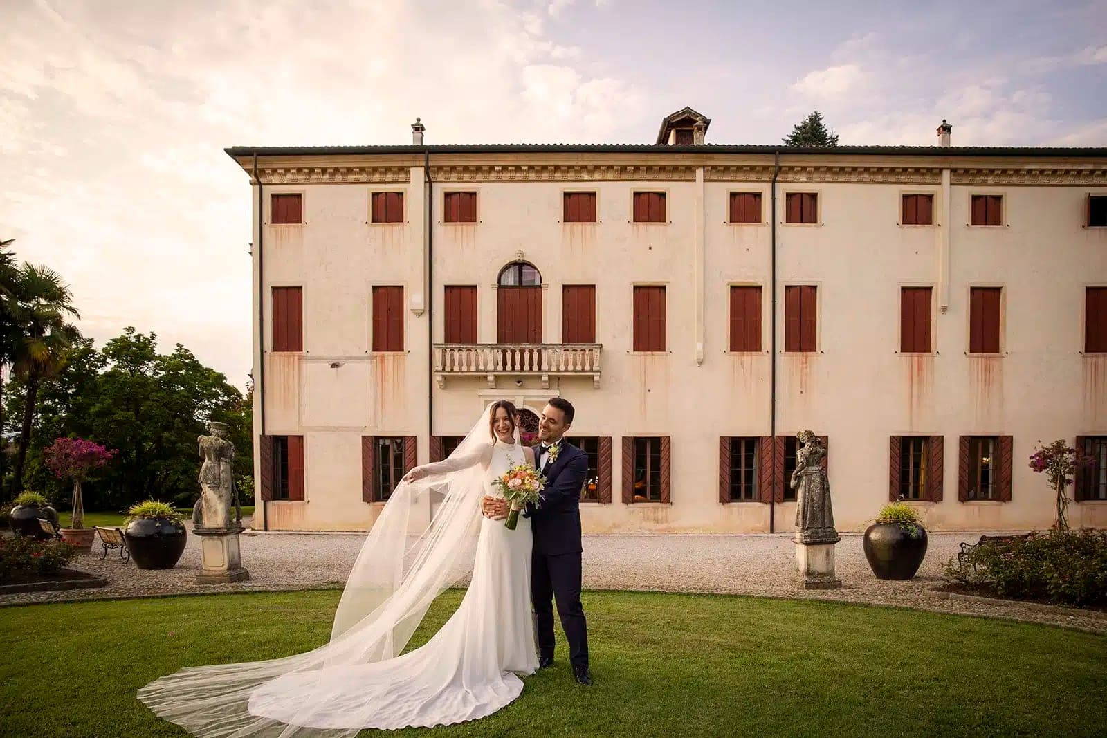 A bride and groom stand on a lawn in front of a large building with red-shuttered windows.