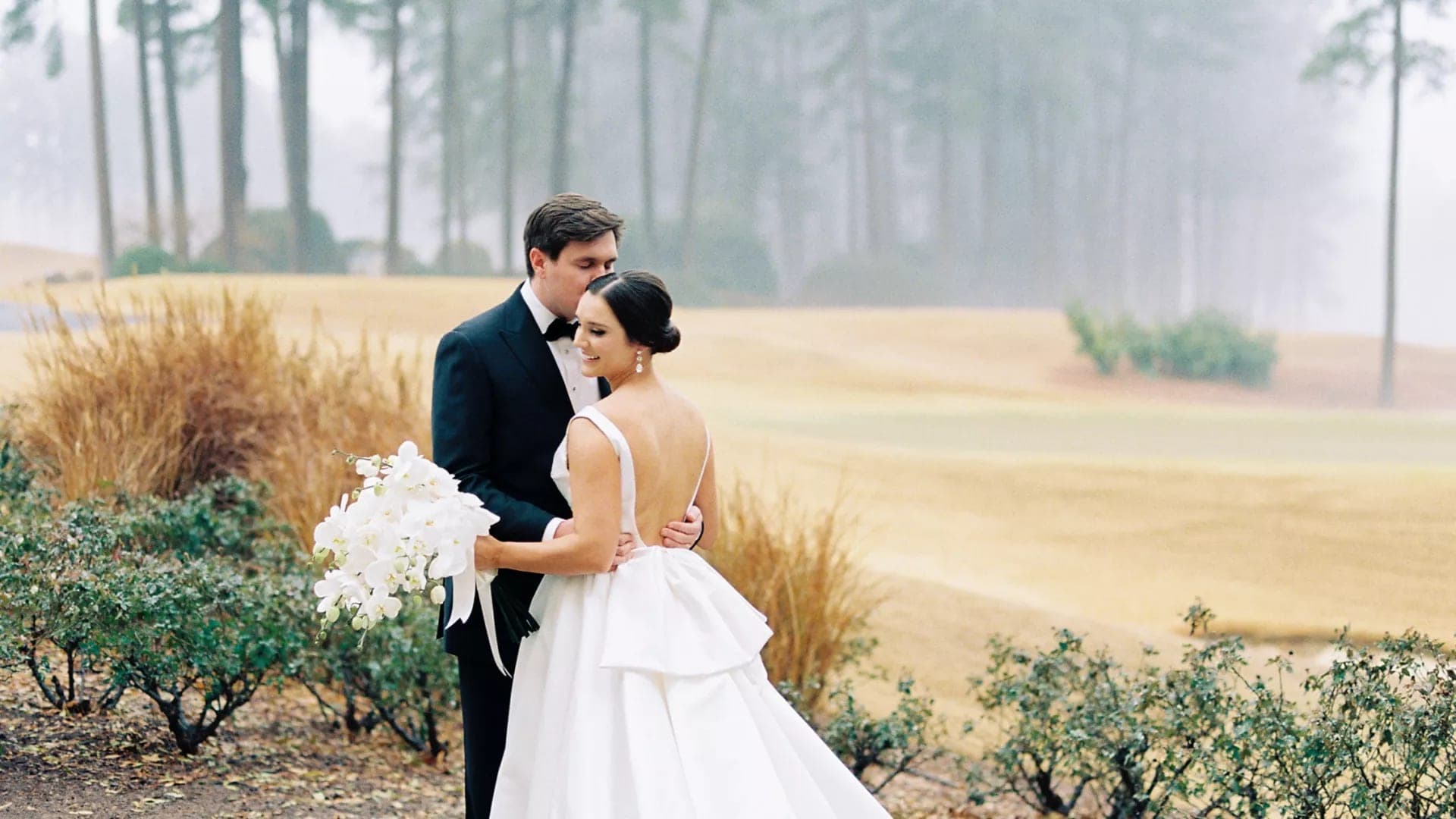 A bride and groom embrace on a golf course with trees in the background.