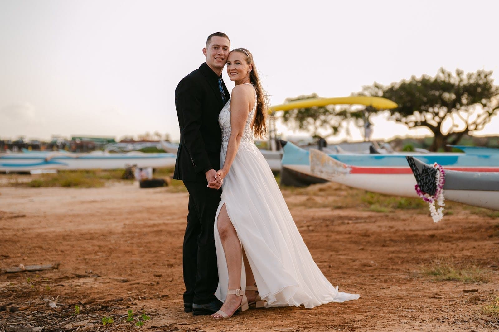 A bride and groom stand holding hands on a beach with canoes behind them.