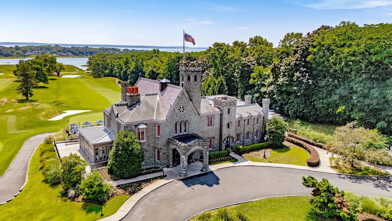 Exterior of a stone castle with a flag on top, surrounded by green lawns and trees, with a body of water in the distance.