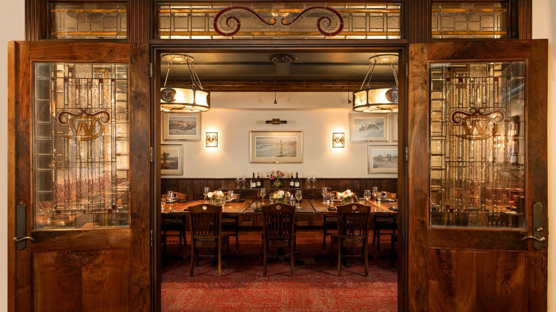 A long wooden dining table set for a meal, viewed through open wooden doors with stained glass.