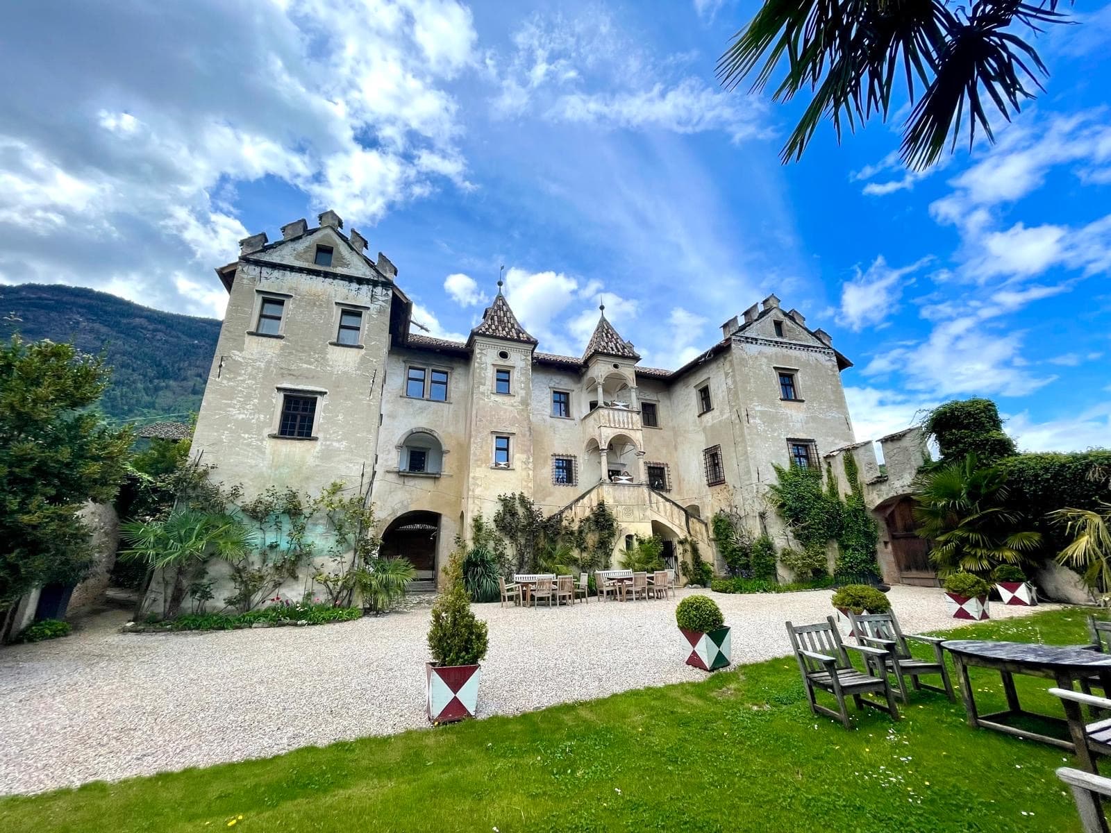 The stone facade of Ansitz Zinnenberg castle with crenellated towers, a gravel courtyard, outdoor wooden chairs, and palm trees under a cloudy sky.