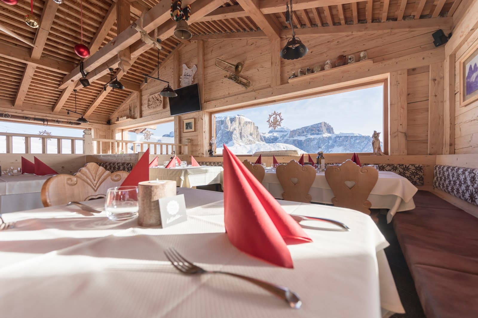 A wood-panelled alpine restaurant dining room with set tables, red folded napkins, and a large window framing a snow-covered rocky peak.